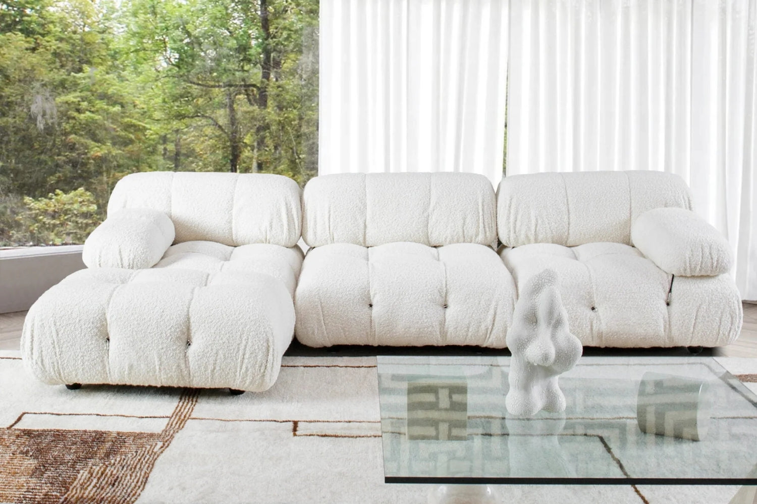 A modern living room with a plush white sofa in front of floor-to-ceiling windows showing a green, leafy outdoor view. A glass coffee table with a white sculptural decoration sits on a patterned rug.