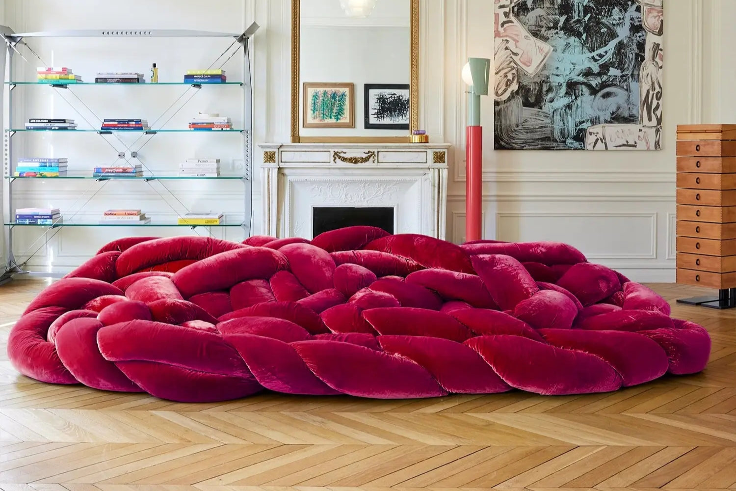 A luxurious living room with a large, braided red velvet sofa in the center. The room features a white fireplace, modern shelving with books, a tall red lamp, and framed artwork on the wall. The floor is wooden with a herringbone pattern.
