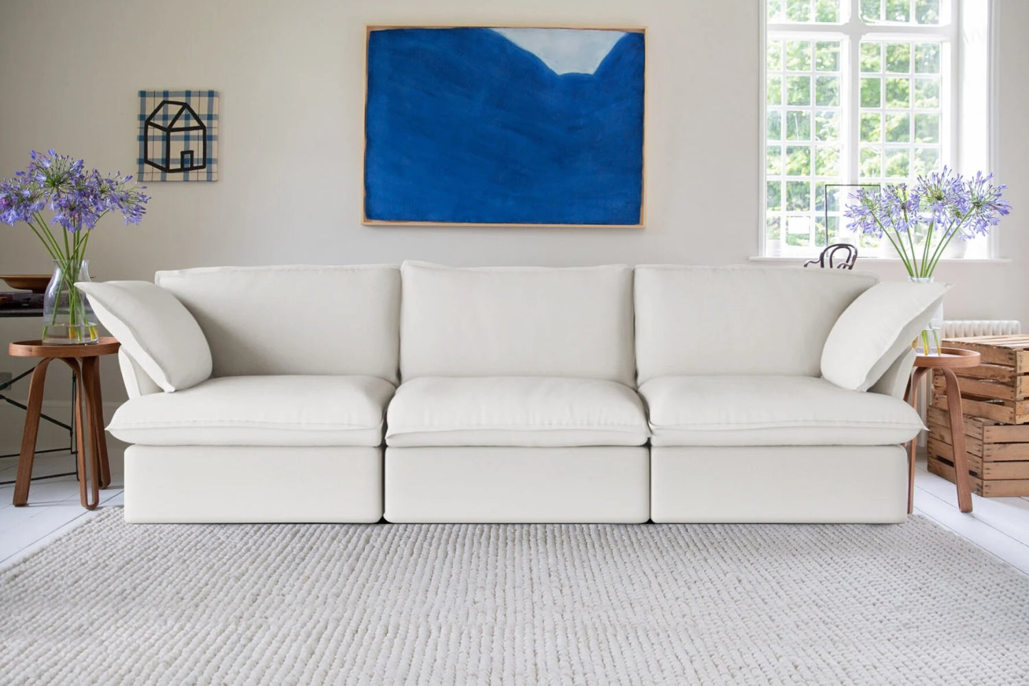 A white sofa with four cushions is in a bright living room. It is flanked by two side tables with vases of purple flowers. A blue abstract painting hangs on the wall above the sofa. A large window lets in natural light.