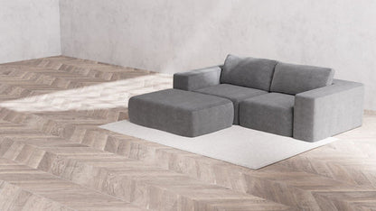 A minimalist room showcasing a light gray Modular 2-Seater Chaise Sectional in Boucle Aspen, complemented by an ottoman, is set against a cream-colored rug. The area boasts herringbone-patterned wooden flooring and light gray walls, establishing a serene and stylish ambiance.