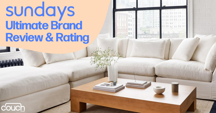 A cozy living room features a large white sectional sofa filled with plush cushions. A light wood coffee table adorned with minimalistic decor, including a small vase and a bowl, sits in front of the sofa. The windows in the background offer a cityscape view. Text on the image reads, "sundays Ultimate Brand Review & Rating." The logo for couch.com is in the bottom left corner.