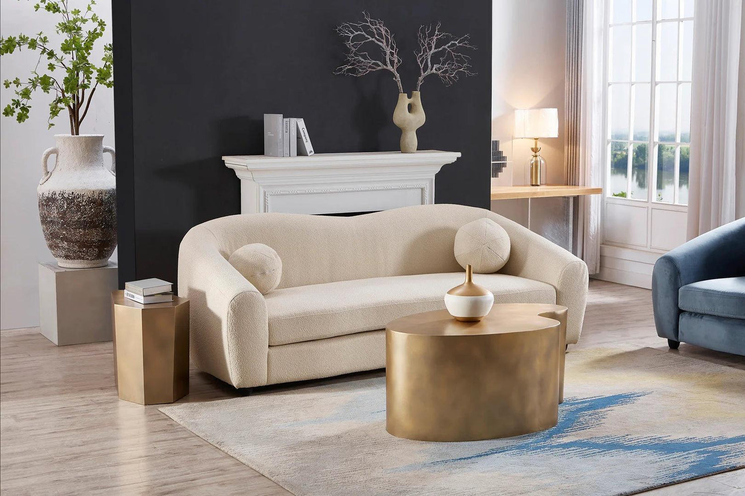A modern living room features a cream-colored sofa with round pillows, a gold coffee table, and a side table with books. Behind the sofa is a fireplace adorned with a decorative vase holding branches. Natural light filters in through large windows, illuminating the space.
