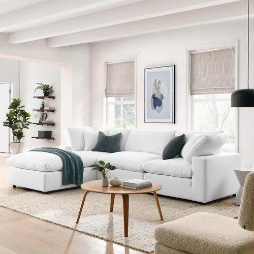 A bright living room with a modern white sectional sofa adorned with gray and white cushions. A green throw blanket is draped over the chaise lounge. A wooden coffee table with a small plant and book sits on a beige rug. Large windows and wall art enhance the space.