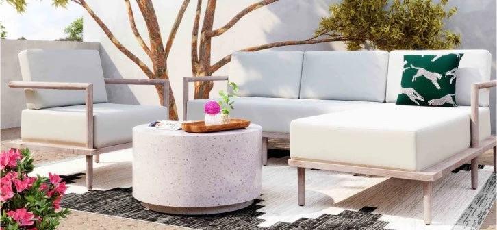 A modern outdoor patio scene featuring light-colored furniture. An armchair, a couch, and a lounge chair surround a circular coffee table with a potted plant. A decorative pillow with a green background and white leopards sits on the couch. Pink flowers adorn the foreground.