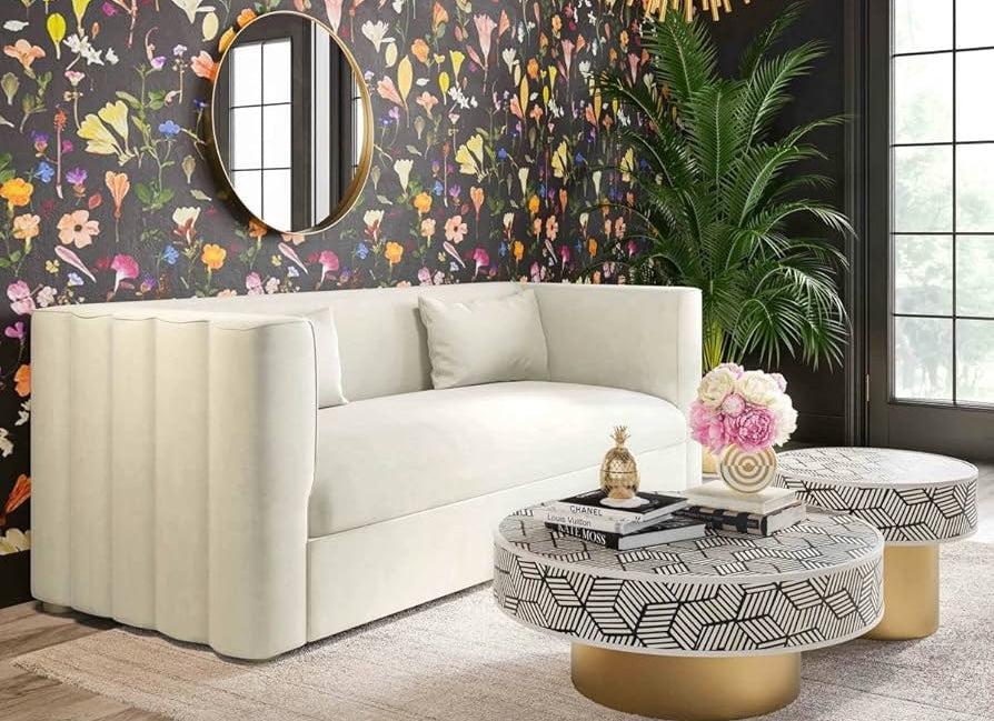 A cozy living room with a white modern sofa against a vibrant floral wallpaper. A large round mirror is above the sofa. Two round coffee tables with geometric patterns and golden bases hold decorative items including a small plant and pink flowers. A tall green plant stands by the window.