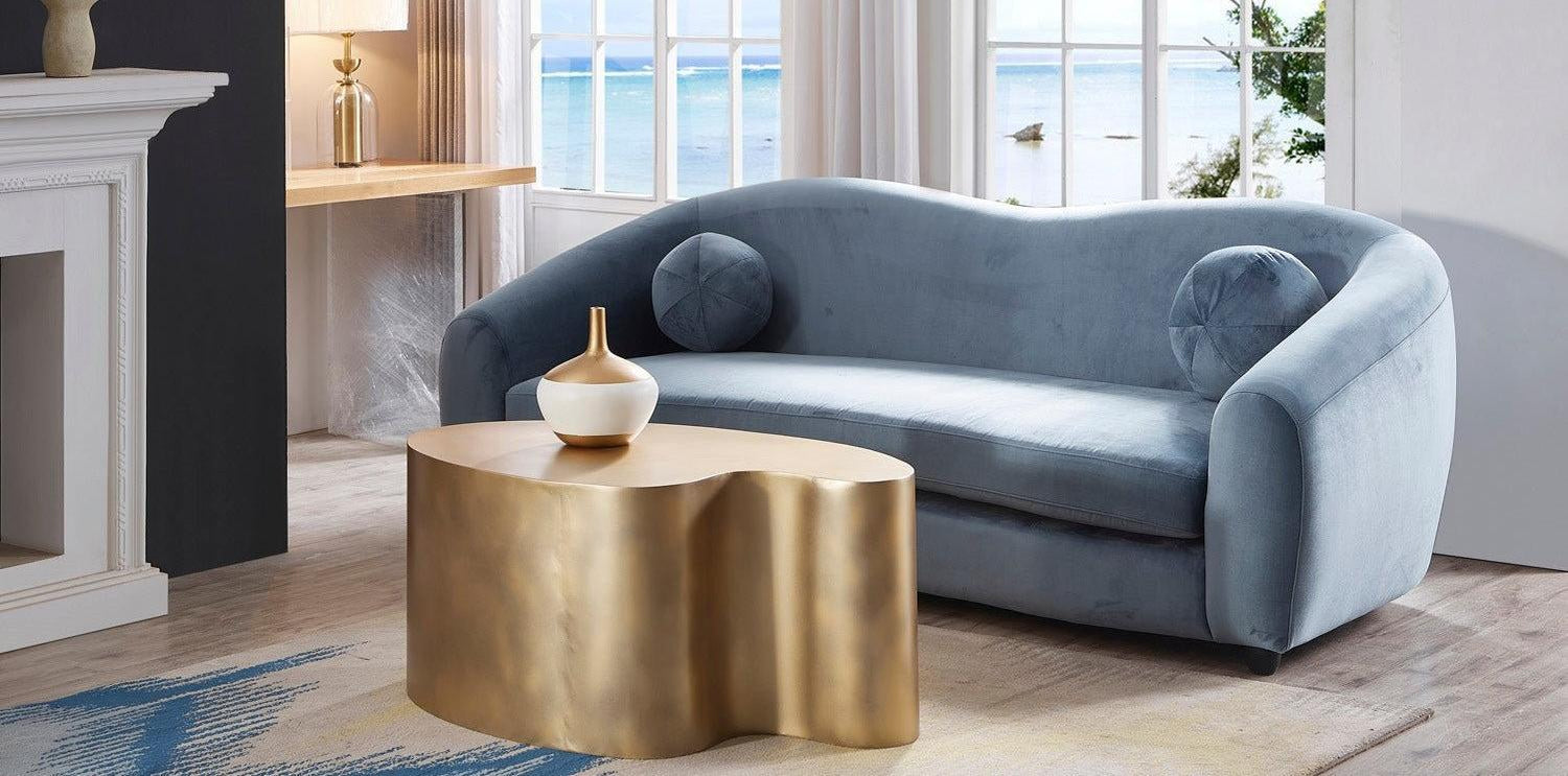 A modern living room features a curvy, blue velvet sofa with circular cushions, a gold, abstract-shaped coffee table, and a scenic view of the ocean through large windows. The space is elegantly decorated with a white vase on the coffee table and a light-colored rug.