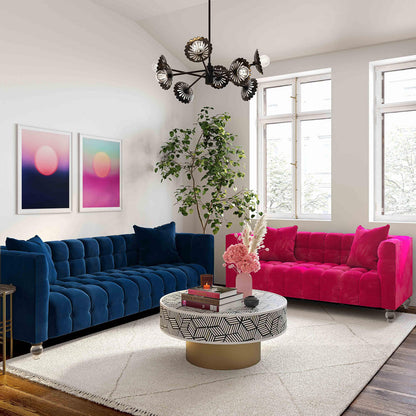 A modern living room features a Bea Velvet Tufted Sofa in blue and another in pink, positioned to face each other. A round coffee table adorned with books and pink flowers rests on a beige rug. On the wall hang two abstract artworks, while a large plant stands by the window.