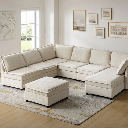 A spacious living room showcases the Victoria Oversized Soft Sectional Sofa with Storage, arranged in an L-shape. A matching ottoman complements it, while a light-colored rug adorns the floor and wall art hangs above the sofa. Large windows provide a beautiful view of the greenery outside.