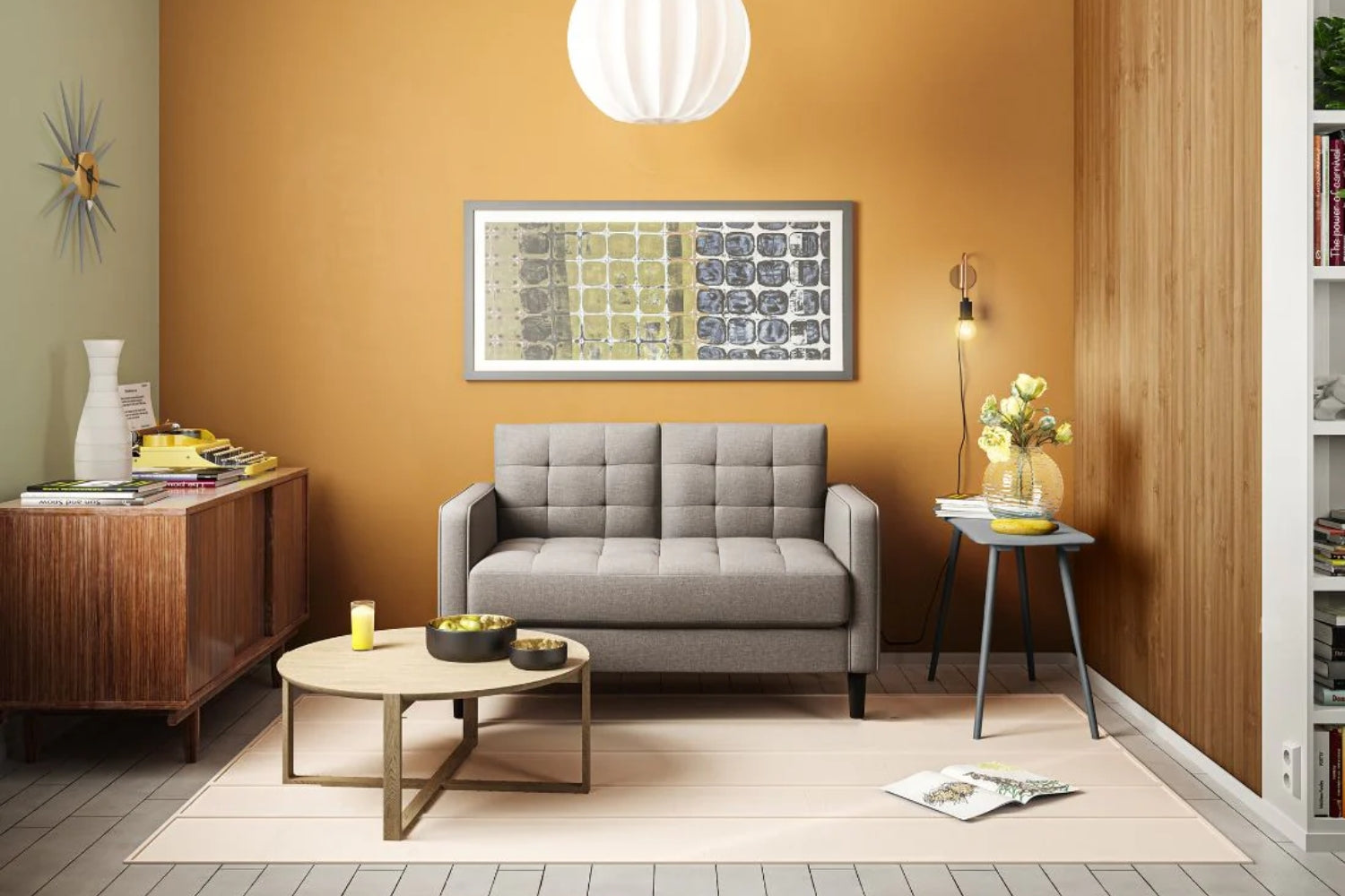 A cozy living room with a gray sofa against an orange accent wall. A round coffee table is centered on a light rug, holding candles. A side table with flowers and books stands to the right. A framed abstract artwork hangs above the sofa.