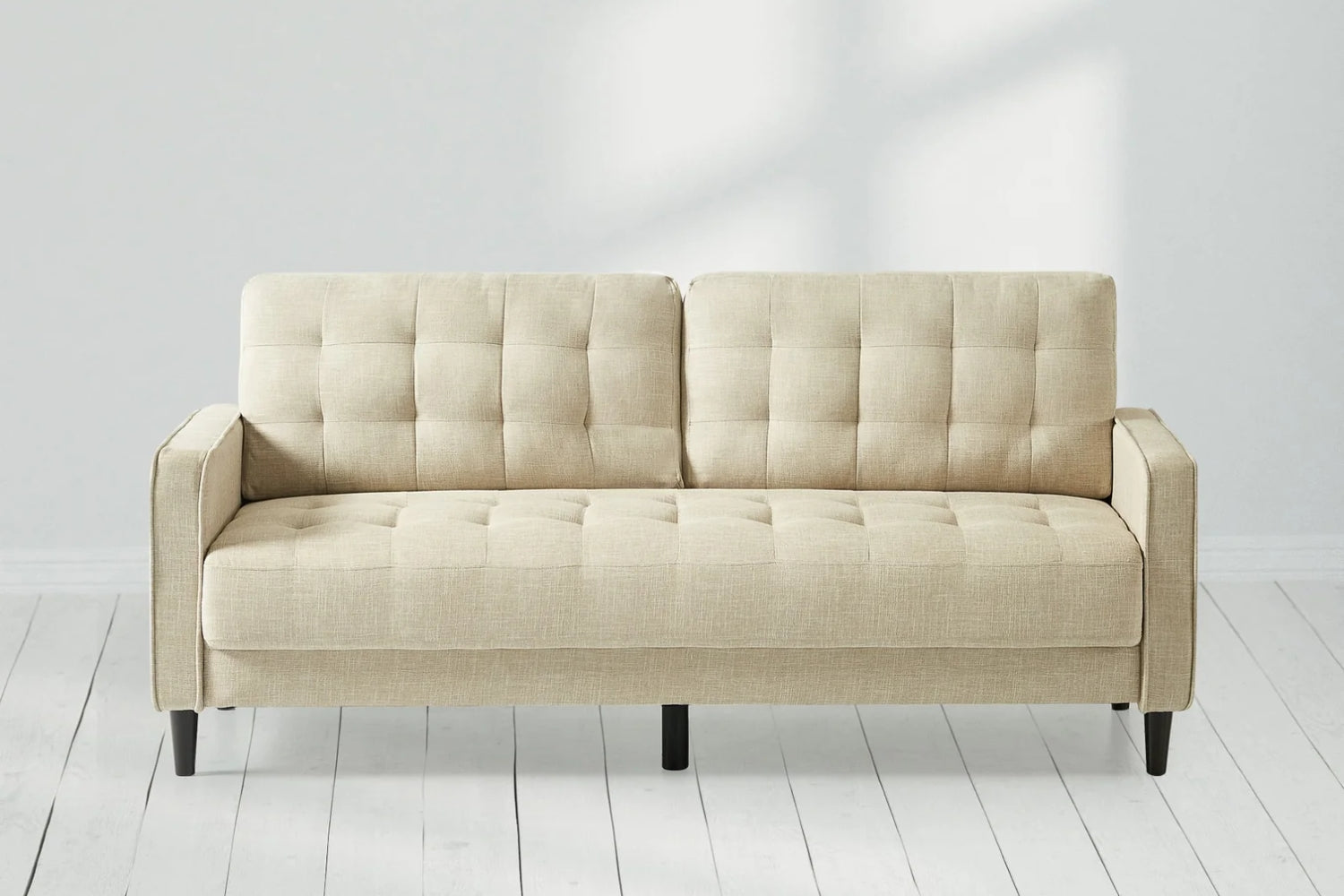 Beige, tufted sofa with a minimalist design set against a light gray wall and placed on a white wooden floor. Sunlight casts soft shadows, enhancing the cozy ambiance.