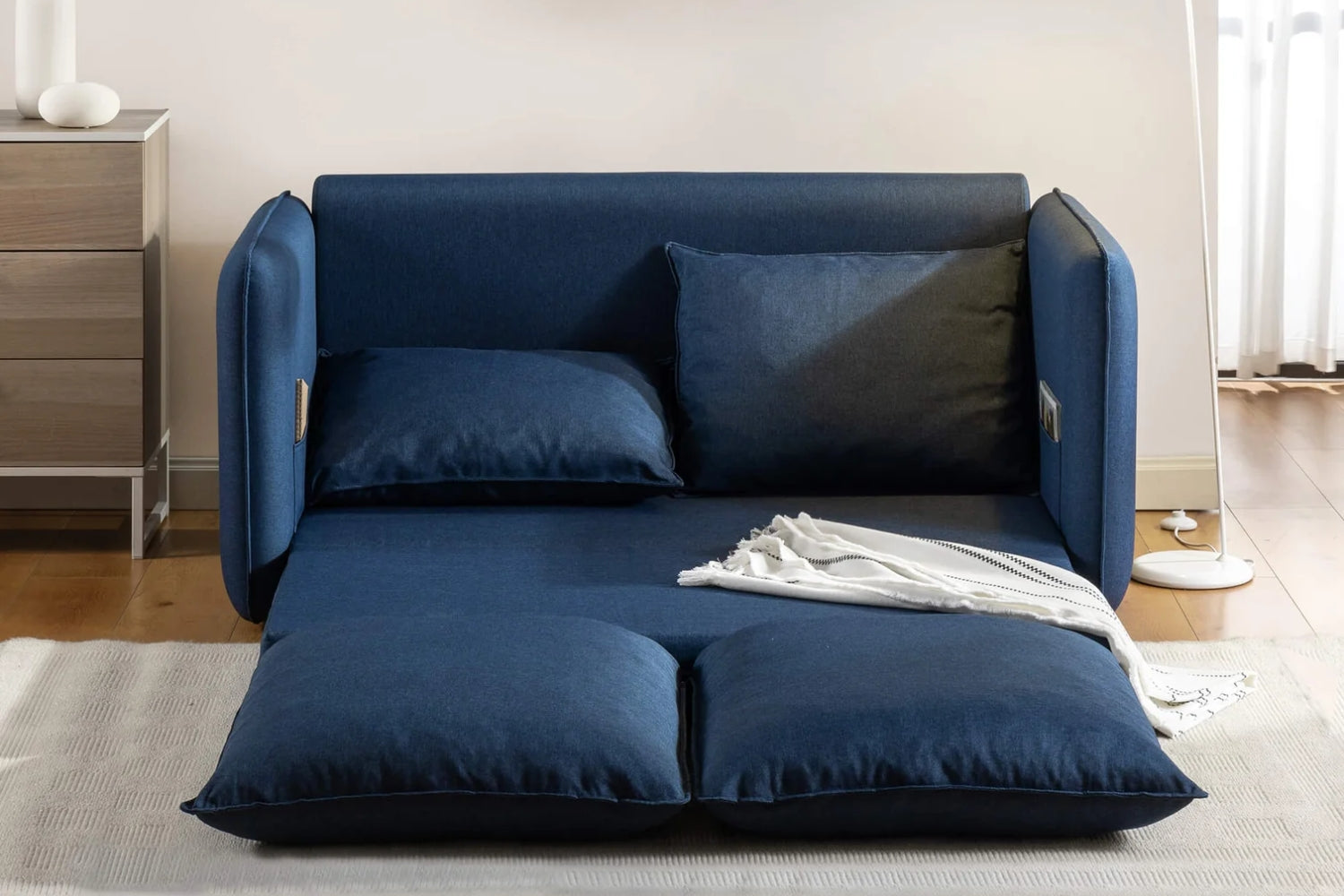 A blue futon sofa bed is in a well-lit room. It is unfolded into a flat position, with two large cushions and a small white blanket on top. A wooden nightstand and a cylindrical lamp are visible in the background.