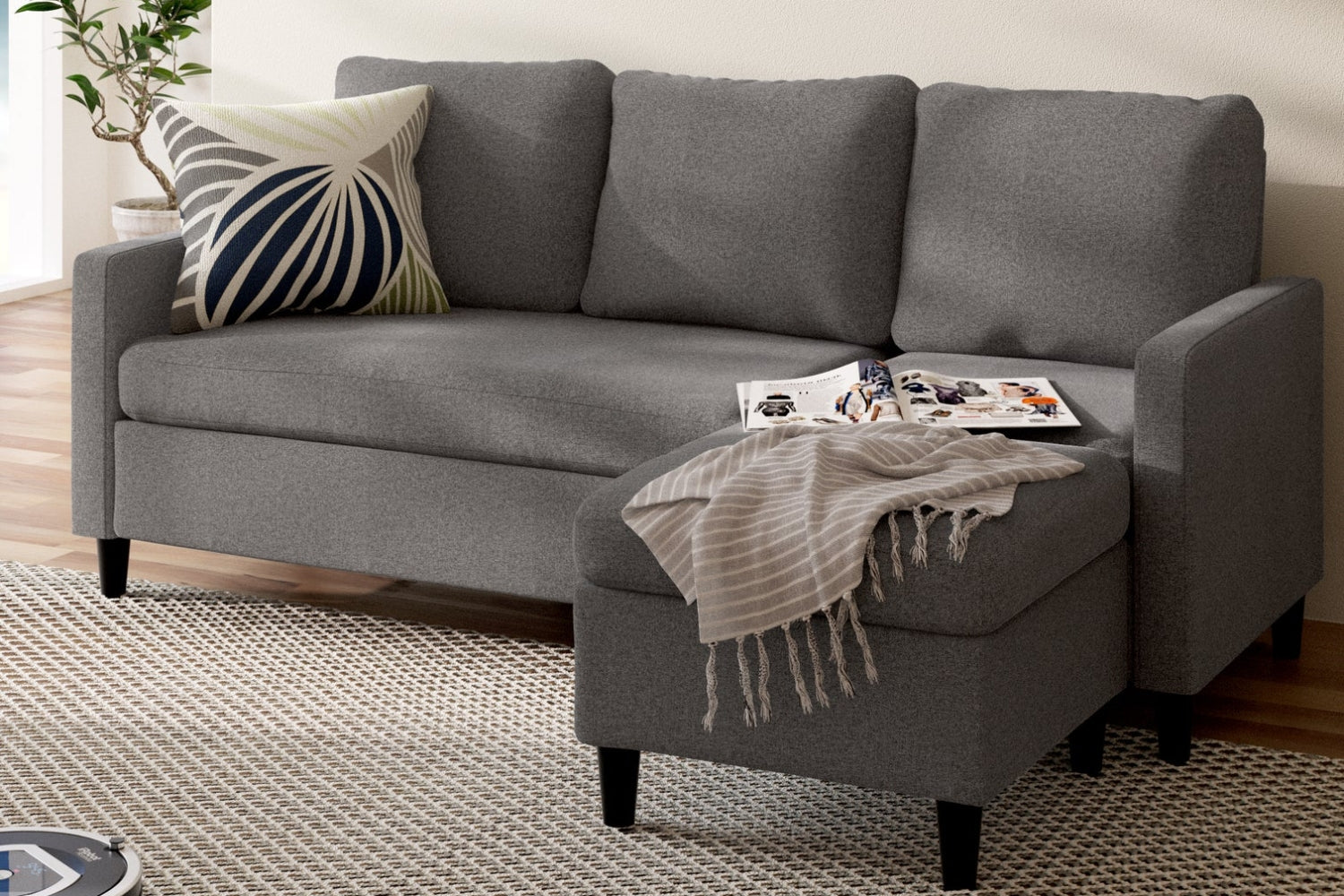 Gray sectional sofa with a chaise lounge in a living room. A patterned throw pillow rests on the left side, and a striped blanket is draped over the chaise. An open magazine lies nearby. The area rug is textured in beige tones.