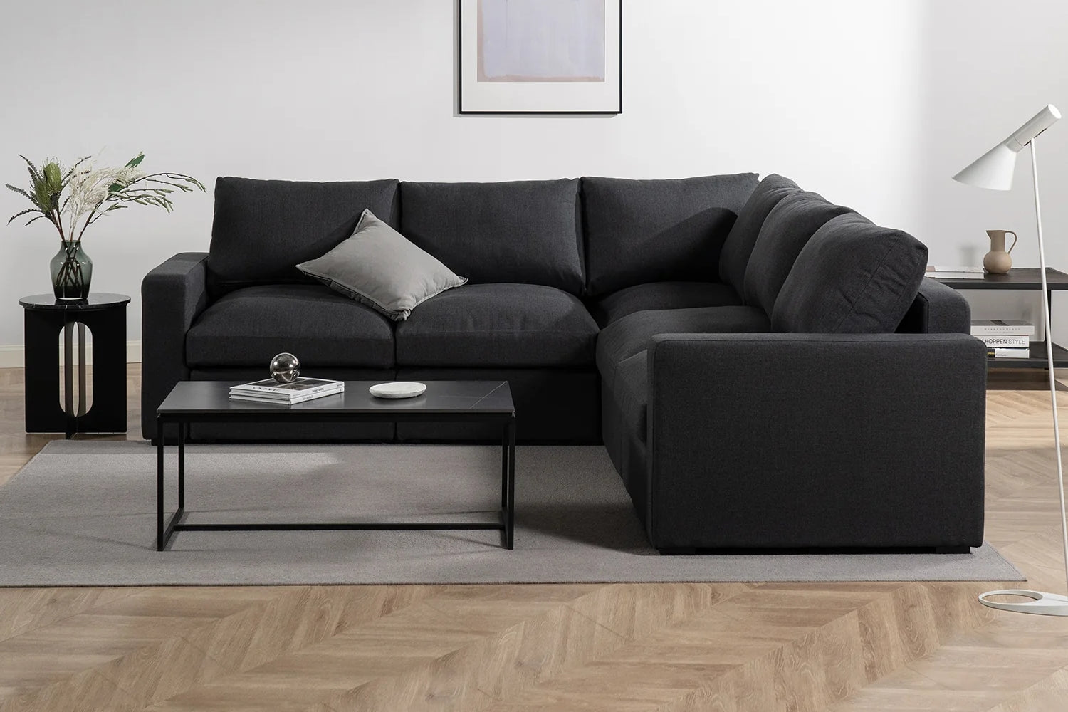 A modern living room with a dark gray sectional sofa, a rectangular coffee table, and a floor lamp. A vase with flowers sits on a side table to the left, and a framed picture hangs on the white wall above the sofa. Light wood flooring is visible.