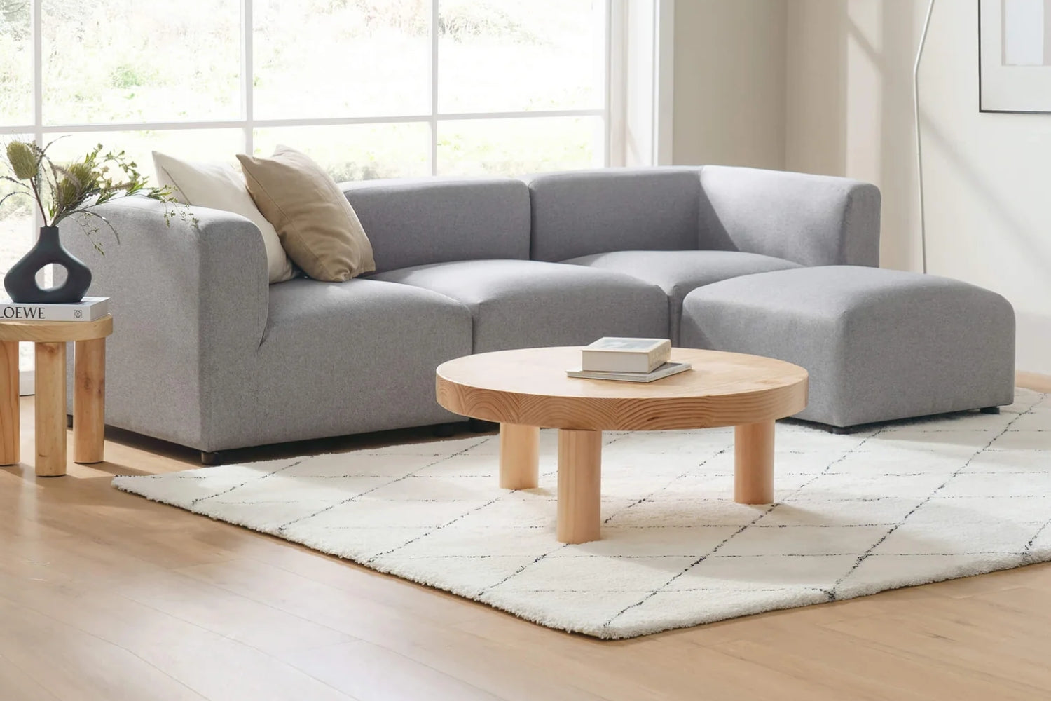 Living room with a modern gray sectional sofa, beige pillow, matching ottoman, round wooden coffee table, small side table with a vase, and a geometric-patterned white rug. Large window with natural light in the background.