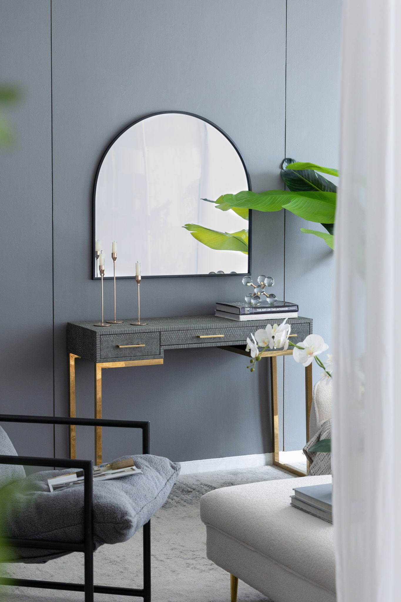 An Arched Mirror With Metal Frame is mounted on a gray wall above a modern vanity with gold legs and a gray tabletop, decorated with candles, books, and an orchid. Green plants and neutral furniture complete the setting.