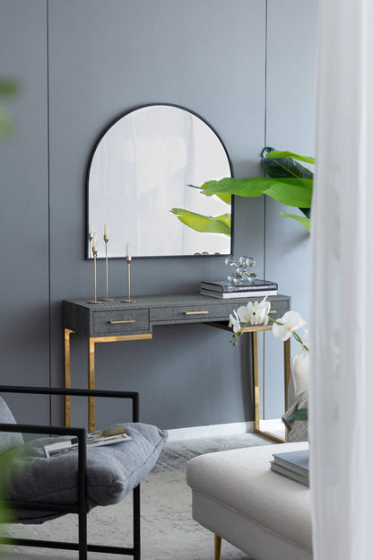 An Arched Mirror With Metal Frame is mounted on a gray wall above a modern vanity with gold legs and a gray tabletop, decorated with candles, books, and an orchid. Green plants and neutral furniture complete the setting.