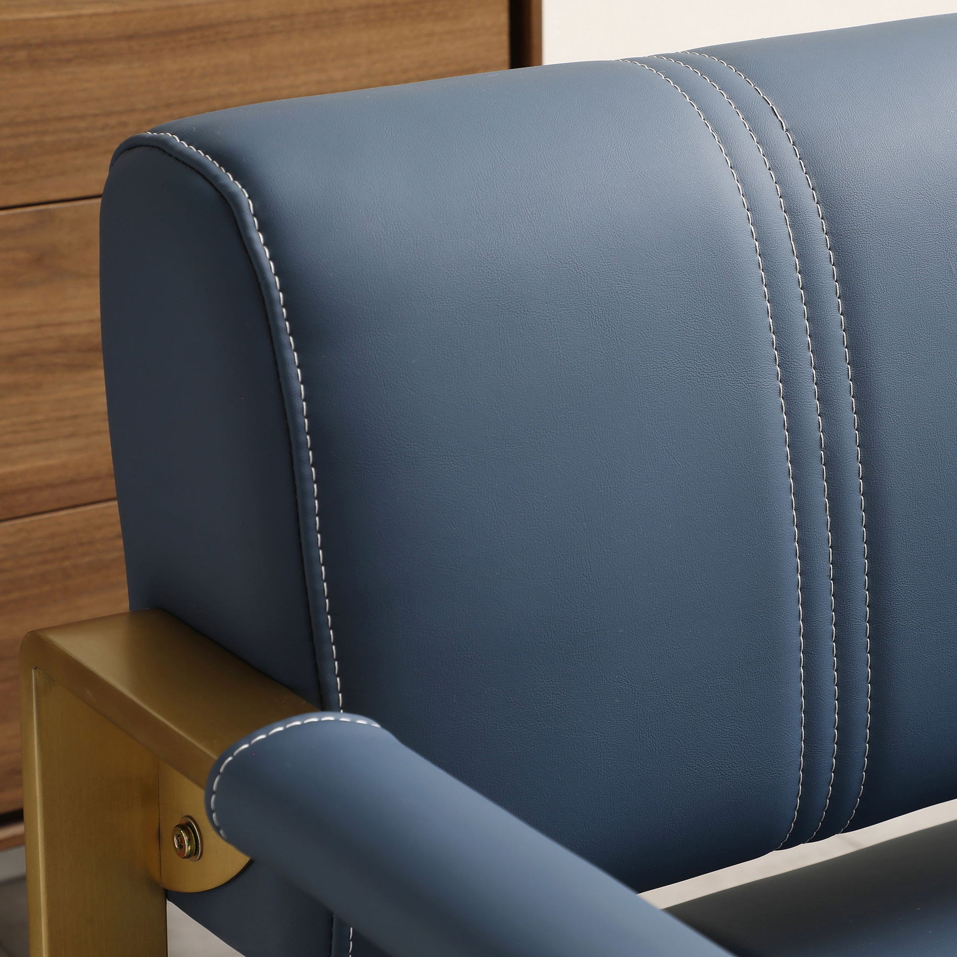 Close-up of the Modern Upholstery Accent Loveseat Sofa, featuring blue leather, white stitching, and golden metal arms. A wooden drawer unit is visible in the background.