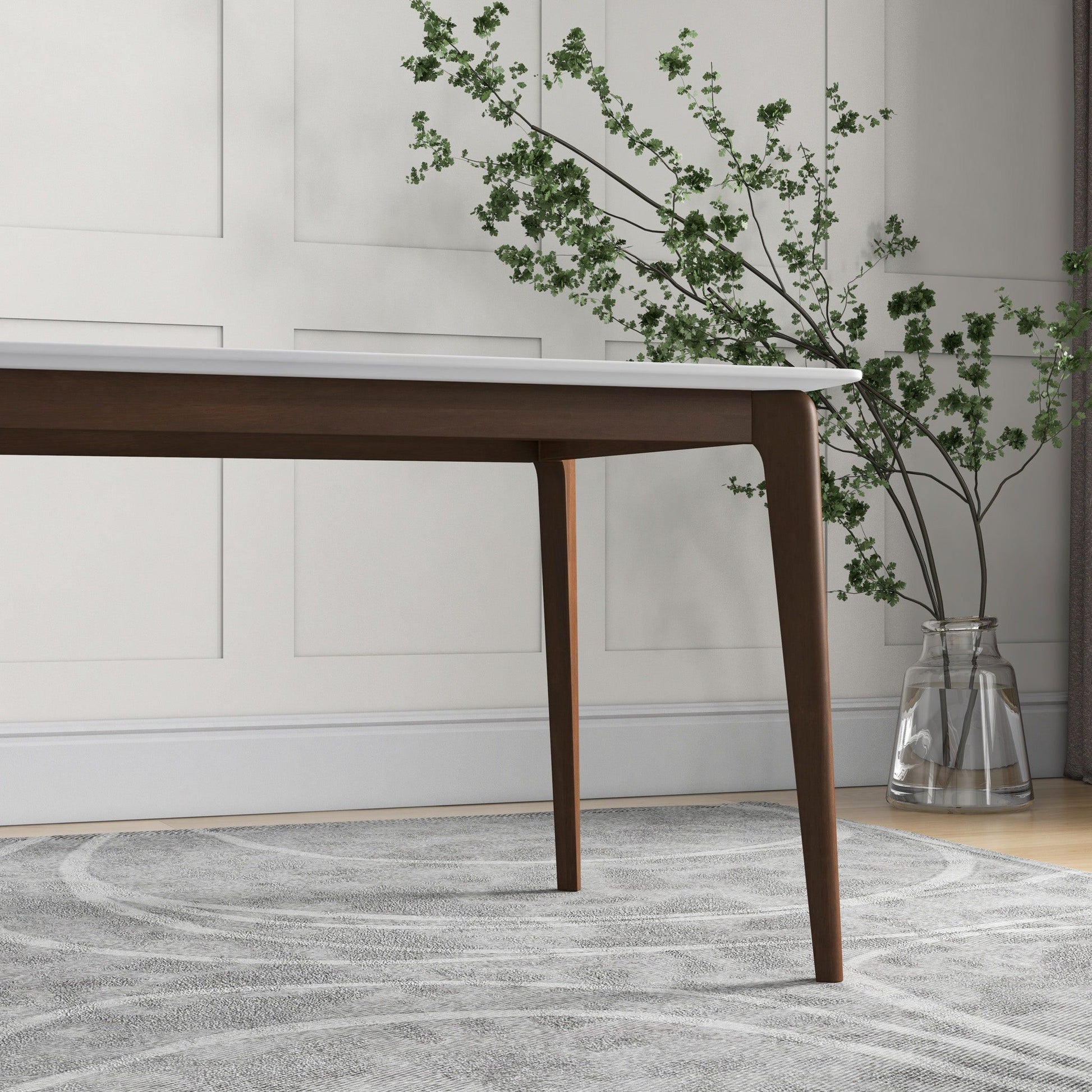The Carlos Dining Table, featuring dark wooden legs and a white top, stands on a light patterned rug. A glass vase with green leafy branches is set against a white paneled wall in the background.