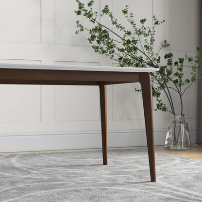 The Carlos Dining Table, featuring dark wooden legs and a white top, stands on a light patterned rug. A glass vase with green leafy branches is set against a white paneled wall in the background.
