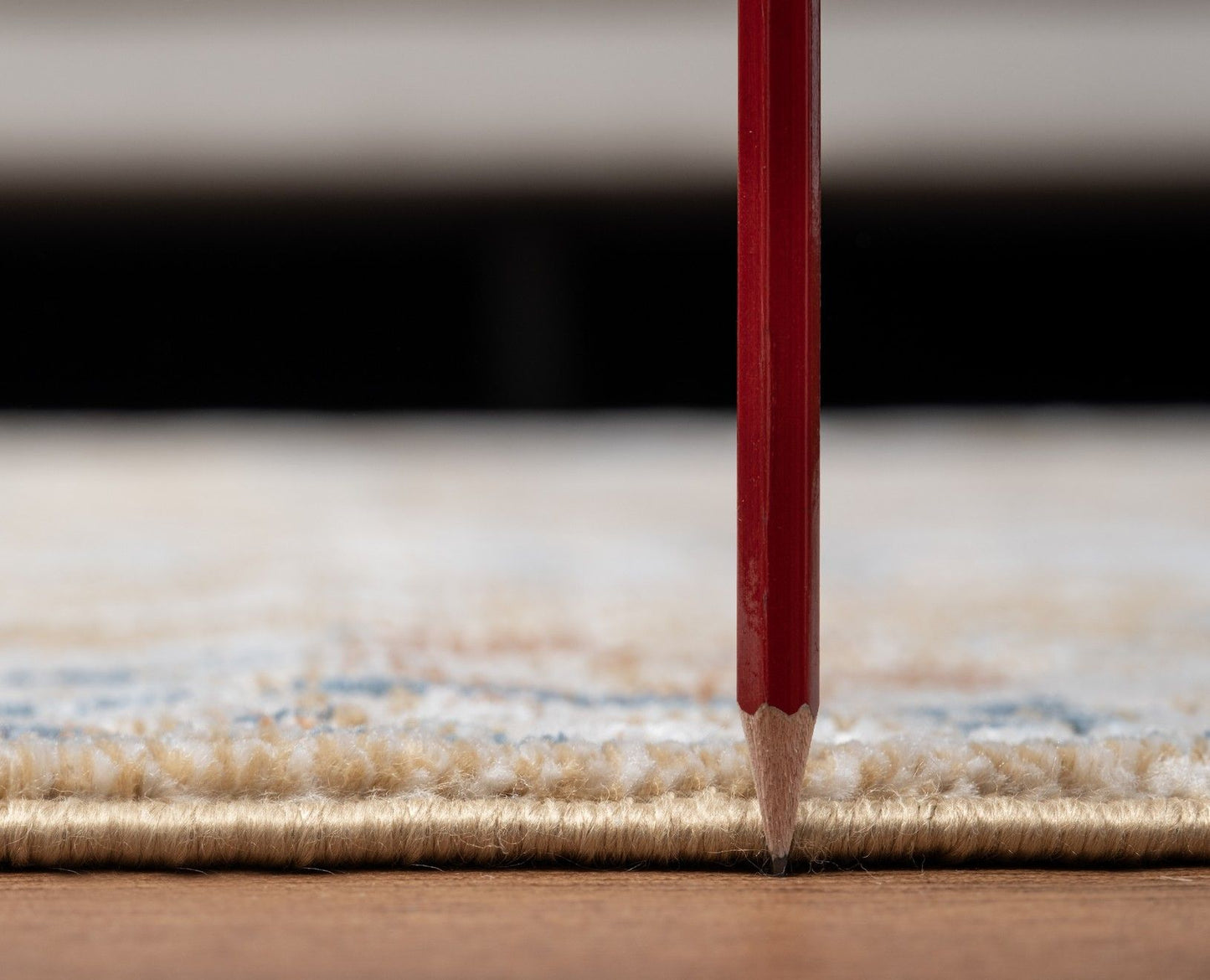 A red pencil stands vertically with its tip touching the edge of the Noble - GC_GEN7008 Area Rug, which rests on a wooden floor; focus is on the pencil and rug edge, with a blurred background.