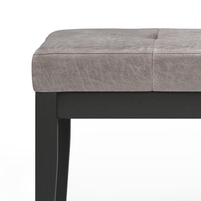 Close-up of the Lacey - Upholstered Tufted Ottoman Bench, featuring a cushioned, light grey textured seat and dark wooden legs against a white background.