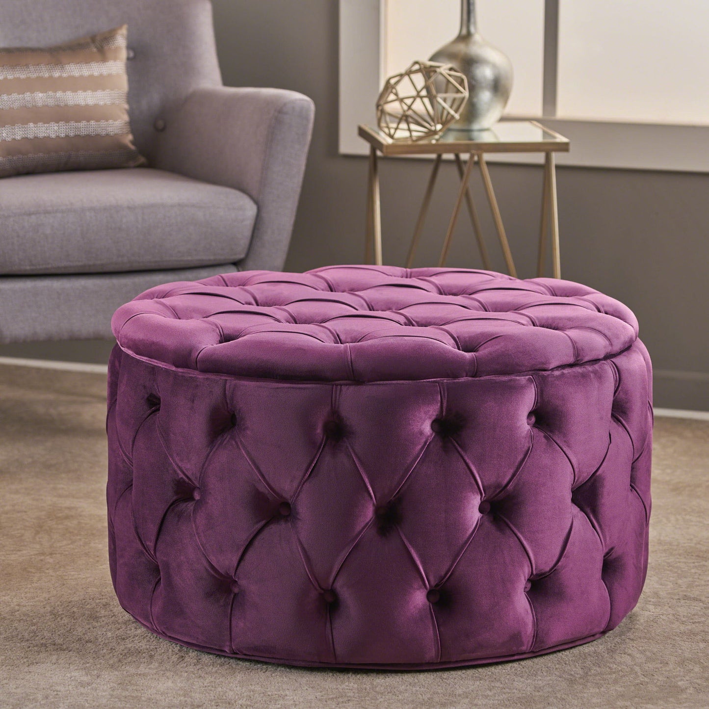The Modern Glam Ottoman Velvet, a round purple tufted piece, sits on a beige carpet in a modern living room with a gray armchair, patterned pillow, and decorated side table in the background.