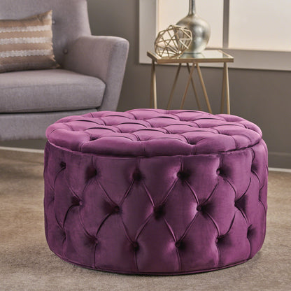 The Modern Glam Ottoman Velvet, a round purple tufted piece, sits on a beige carpet in a modern living room with a gray armchair, patterned pillow, and decorated side table in the background.