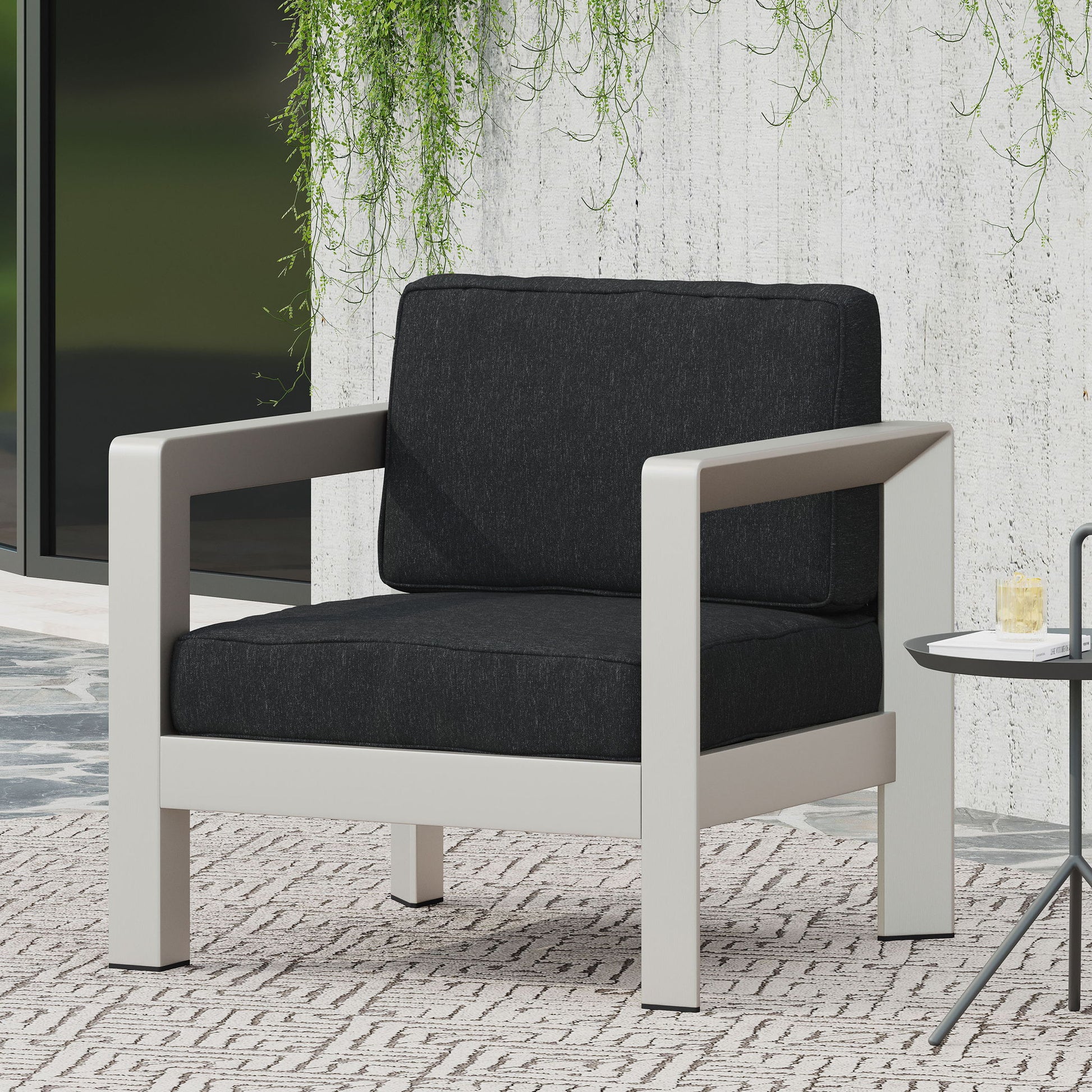 The Aviara - Luxurious Outdoor Club Chair features a light gray metal frame and black cushioned seat and backrest, set on a patterned rug beside a small round table with a drink, against a white textured wall with hanging greenery.