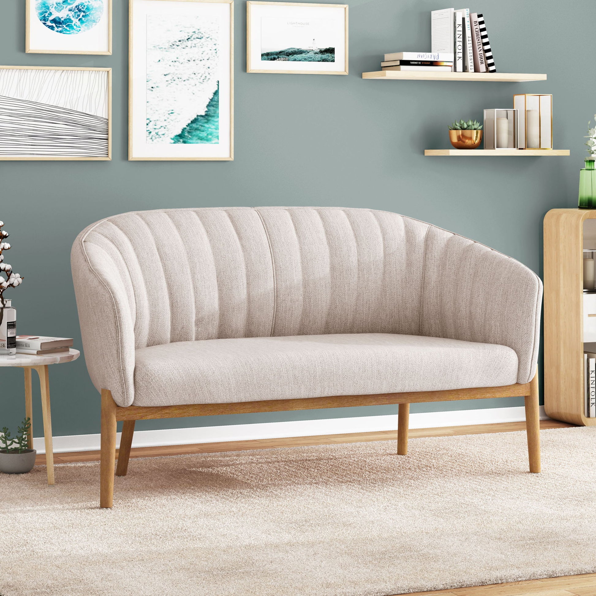 A modern living room showcases the Mid Century Modern Loveseat With Channel Stitched Backrest with wooden legs on a cream rug, while art, books, and plants decorate floating shelves and muted green walls.