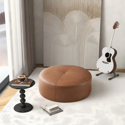 A cozy room featuring the Rochelin Ottoman in tan, a small black side table with a drink and book, abstract wall art, and an acoustic guitar on a stand by a window with sheer curtains.