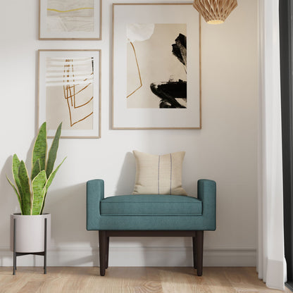 The Scott Upholstered Ottoman Bench in teal, paired with a beige pillow, rests against a white wall beside a potted snake plant. Two abstract art pieces hang above, while a wooden floor and pendant light enhance the modern, minimalist space.