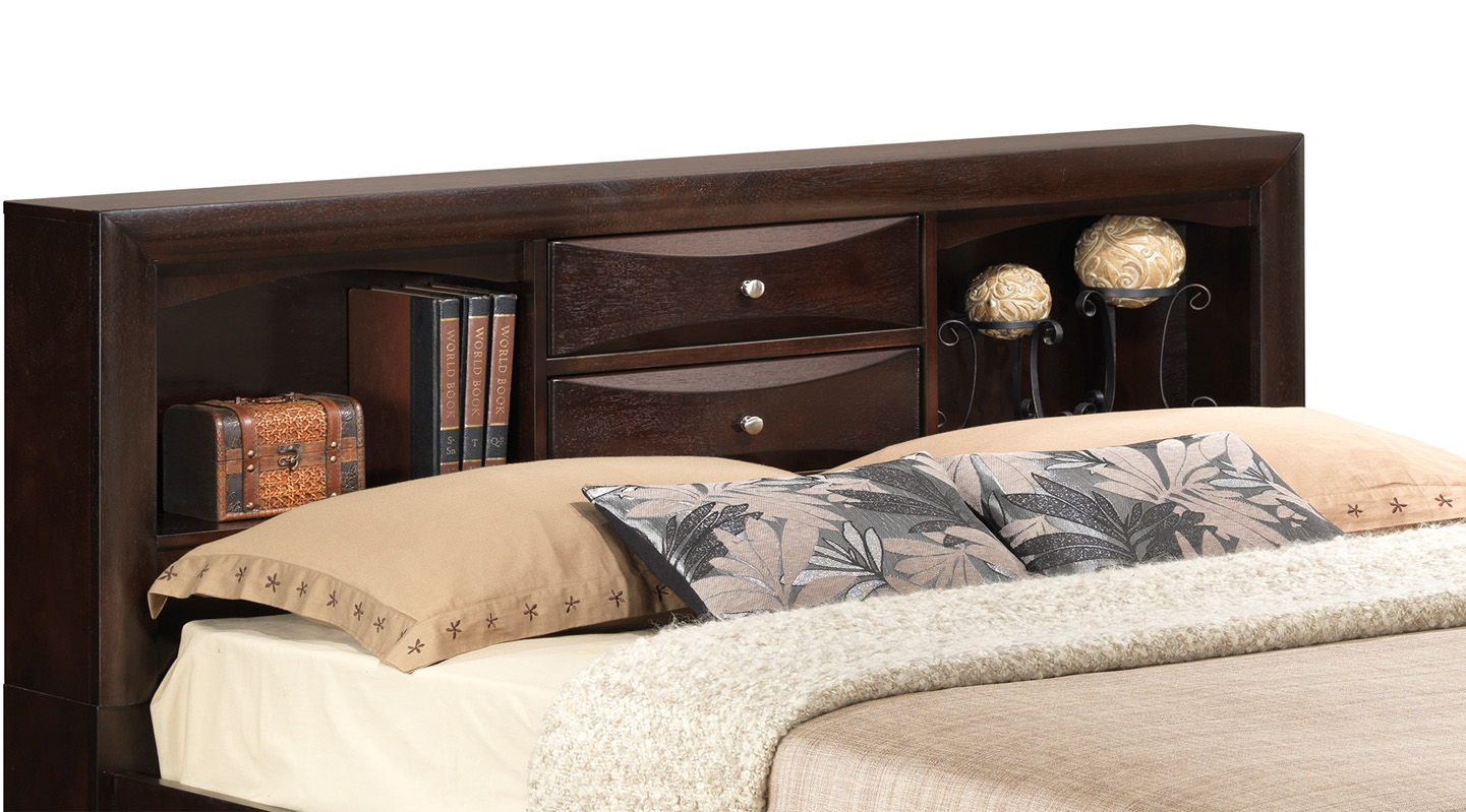 The Marilla Storage Bed With Bookcase Headboard features a dark wood finish, built-in shelves, two small drawers, and displays books, décor, and pillows with floral patterns on a neatly made bed.