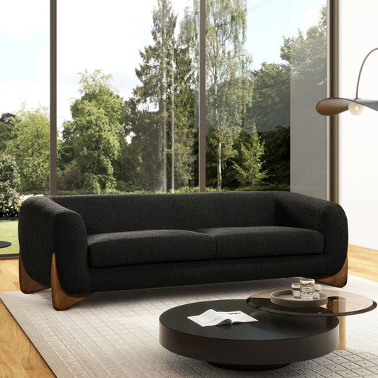 A modern living room featuring the Alpine - Luxurious Upholstery Sherpa Sofa, circular black coffee tables, and a floor lamp on a light rug. Large windows reveal green trees and bright outdoor views.