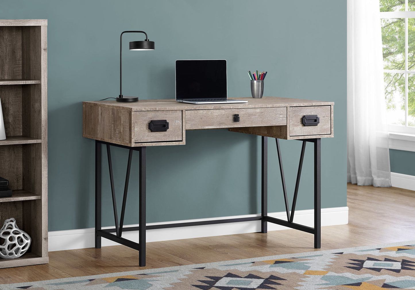 A Computer Desk For Home Office with storage drawers holds a closed laptop, black lamp, and silver cup with pens. To the left is a bookcase; on the floor is a patterned rug; to the right, a window with white curtains.