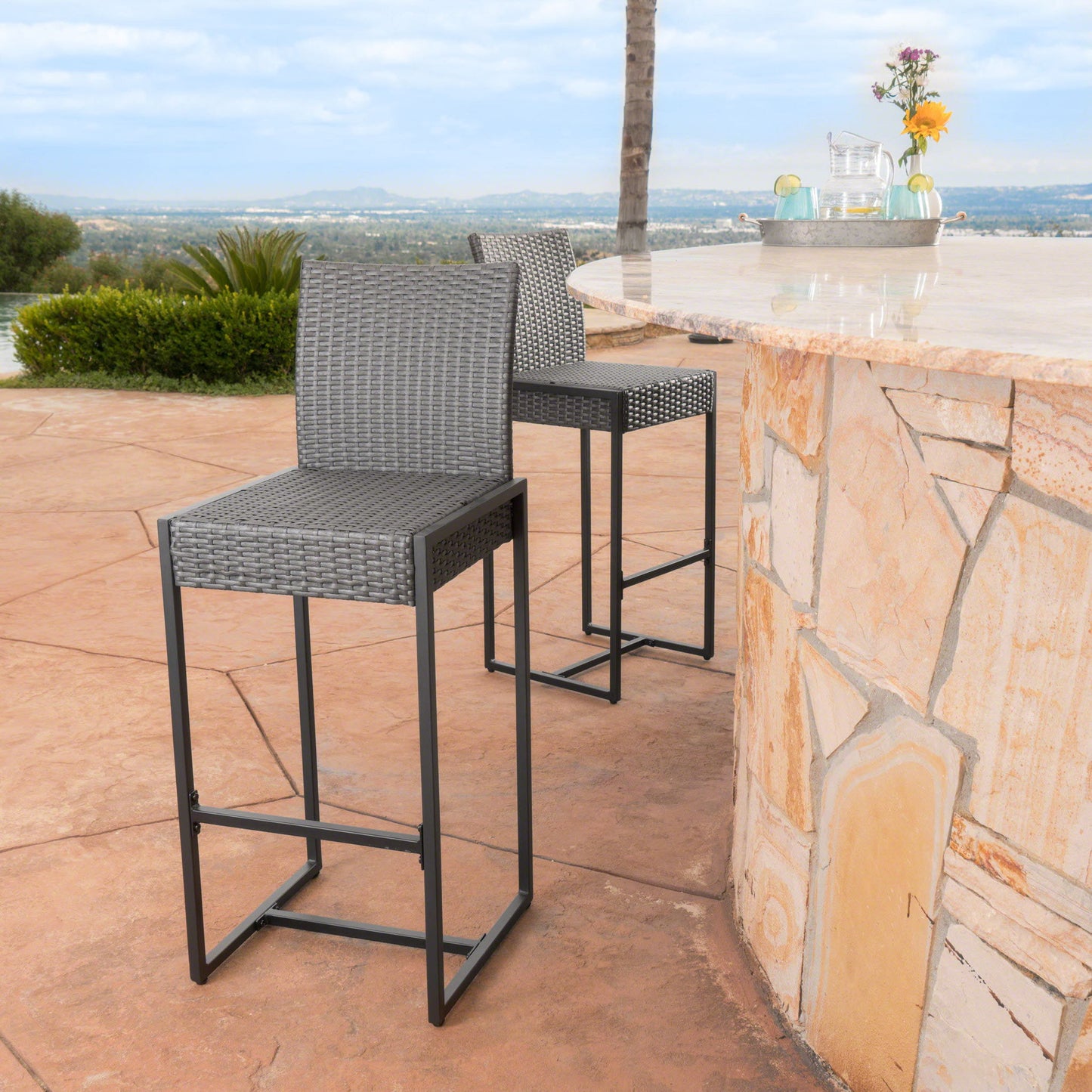A set of two Outdoor Barstools With Hand Crafted Details featuring gray wicker and metal legs are placed by a stone patio bar, overlooking scenic hills and greenery. A vase with flowers decorates the bar.
