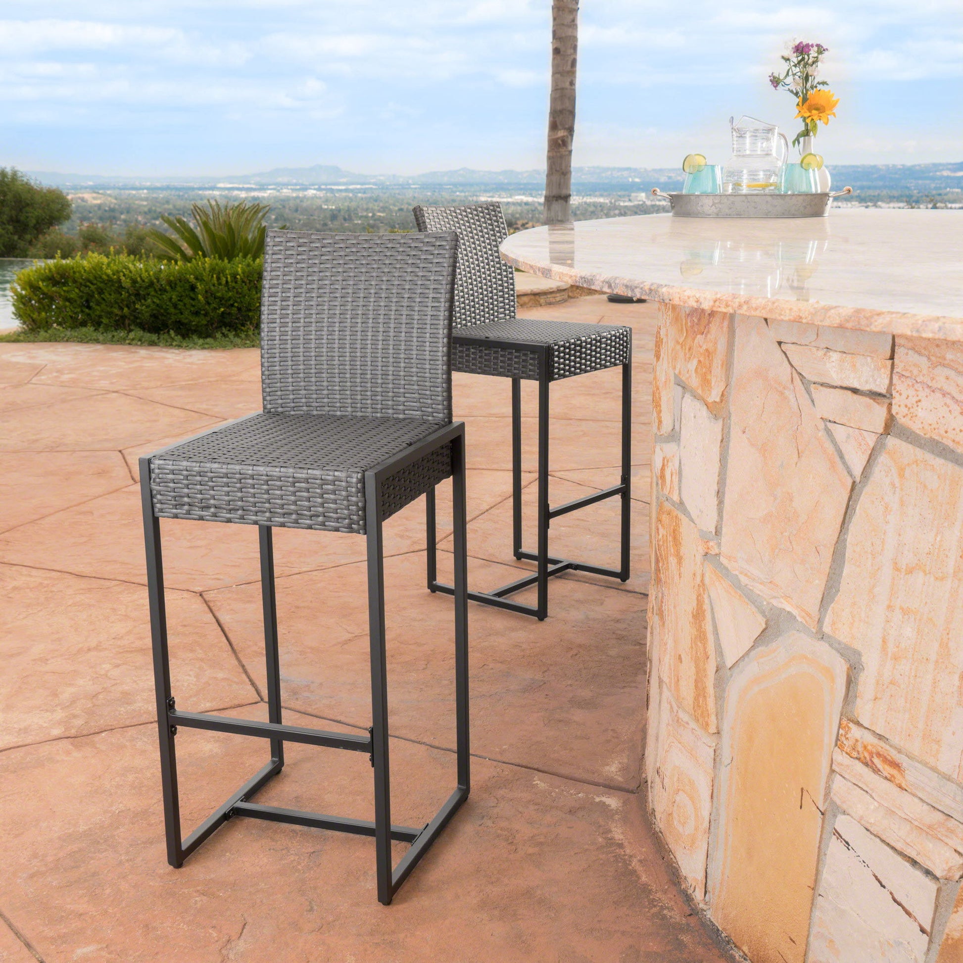 A set of two Outdoor Barstools With Hand Crafted Details featuring gray wicker and metal legs are placed by a stone patio bar, overlooking scenic hills and greenery. A vase with flowers decorates the bar.