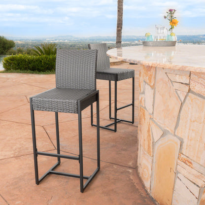 A set of two Outdoor Barstools With Hand Crafted Details featuring gray wicker and metal legs are placed by a stone patio bar, overlooking scenic hills and greenery. A vase with flowers decorates the bar.
