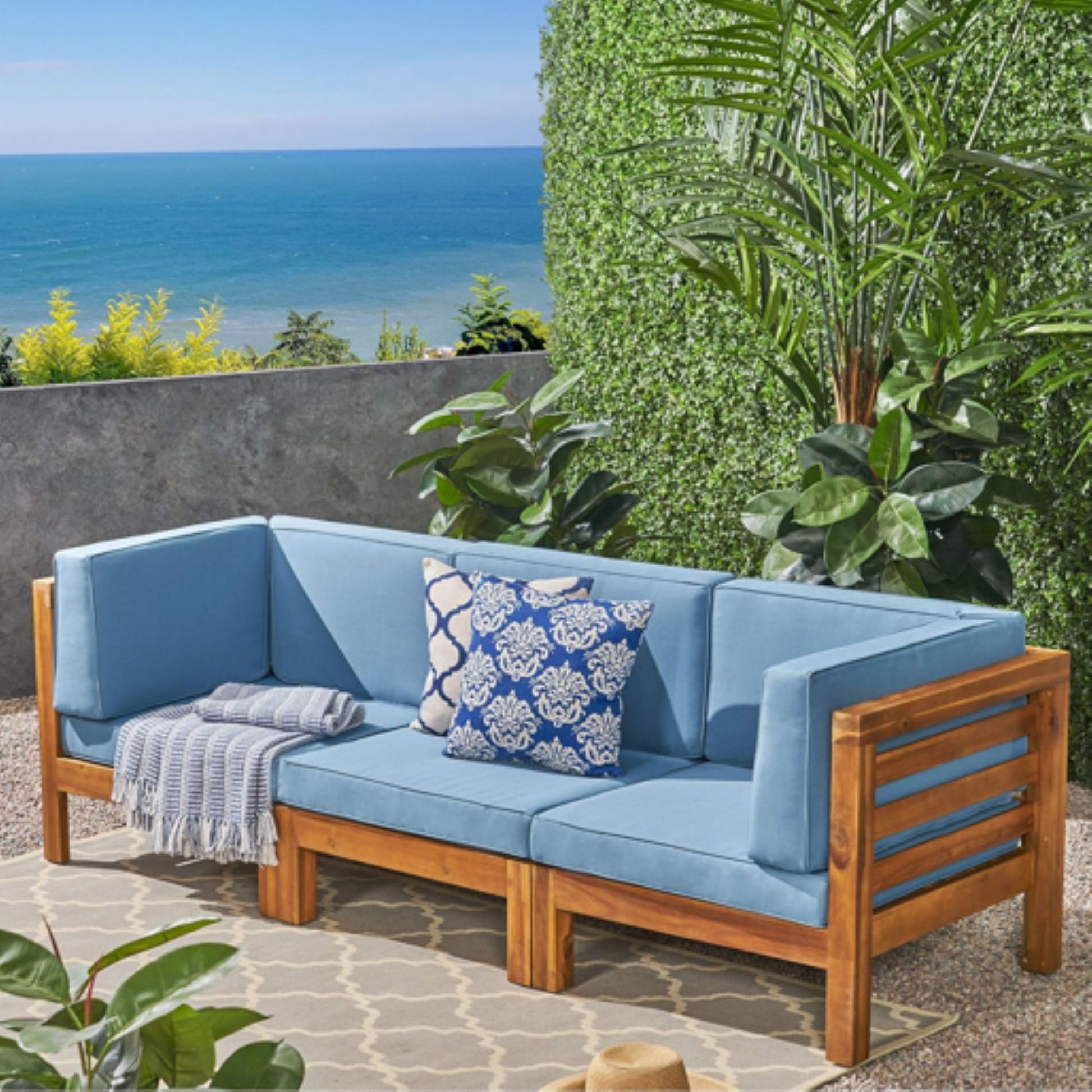 The Oana 3 Piece Sofa Set with water resistant blue cushions is arranged on a patio with a gray and white patterned rug, a blue throw, and a decorative pillow, surrounded by green plants and overlooking the sea.