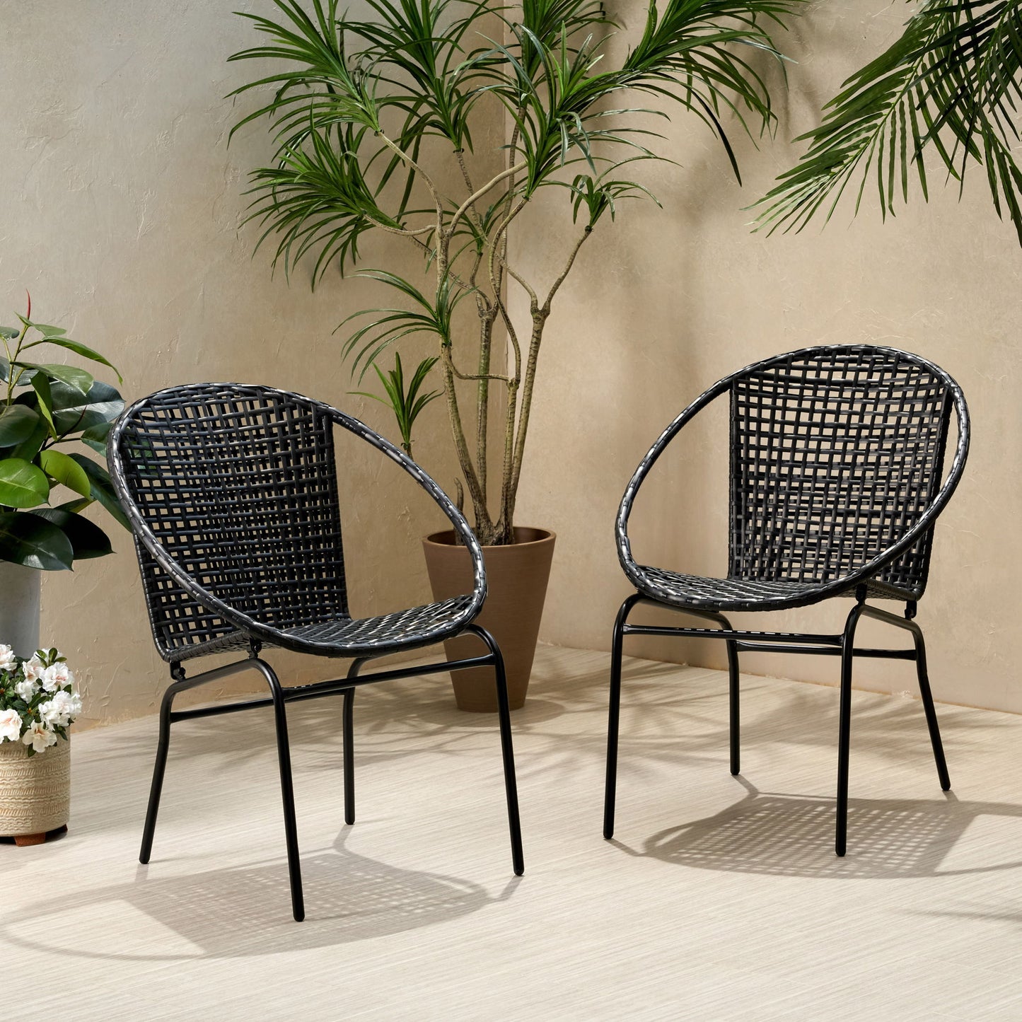 Two Java - Chair With Comfortable Design in black woven finish with metal legs sit on a light floor, surrounded by potted green plants against a beige wall. Soft shadows and natural light create a relaxed, inviting setting.