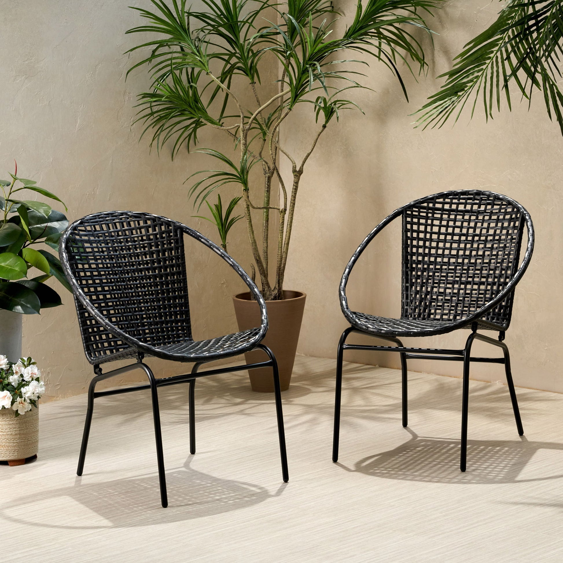Two Java - Chair With Comfortable Design in black woven finish with metal legs sit on a light floor, surrounded by potted green plants against a beige wall. Soft shadows and natural light create a relaxed, inviting setting.