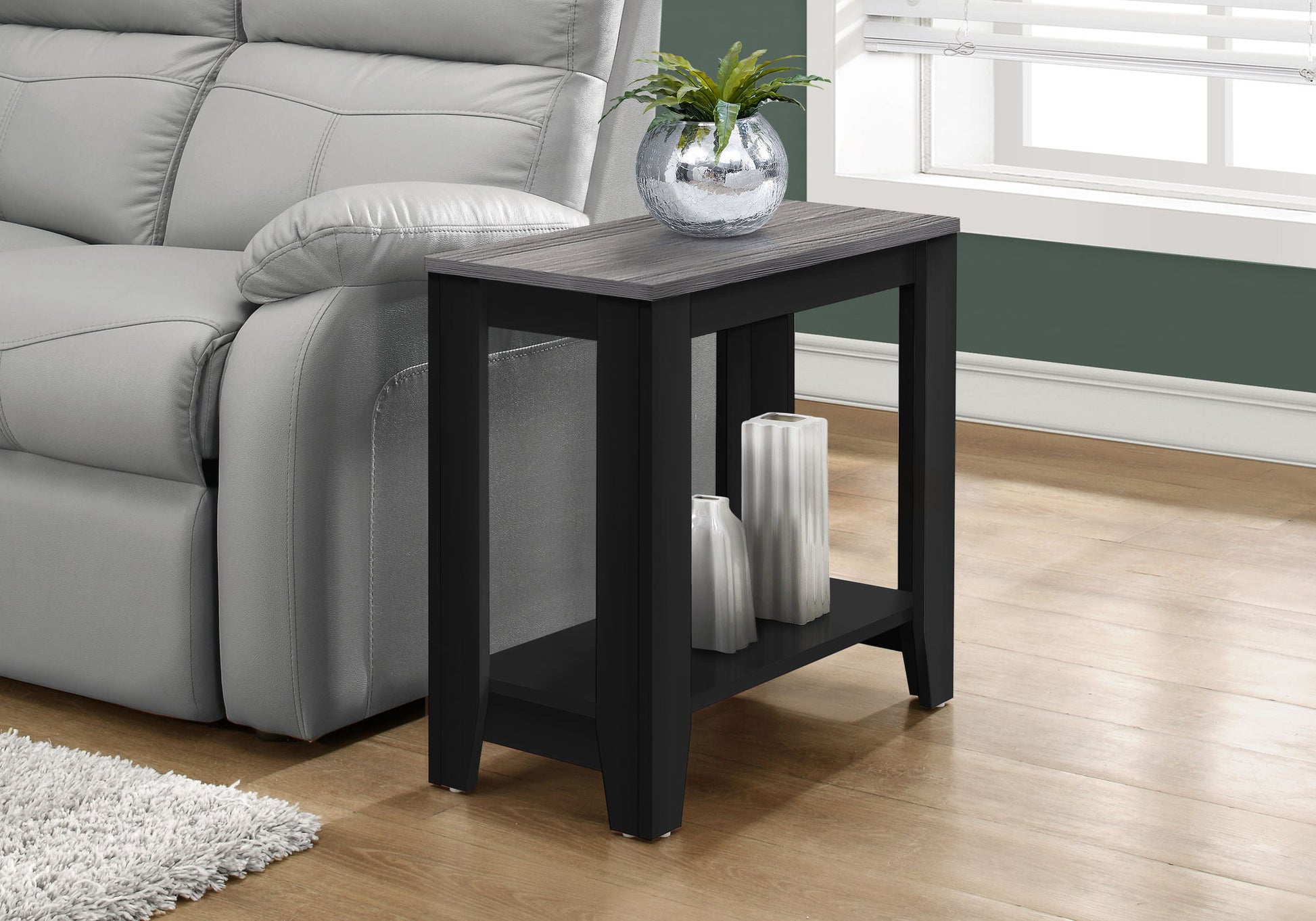 An Accent Table, Side Transitional with elegant design in black and gray stands by a gray sofa. It displays a round glass vase with a green plant on top and two white decorative vases on its lower shelf.