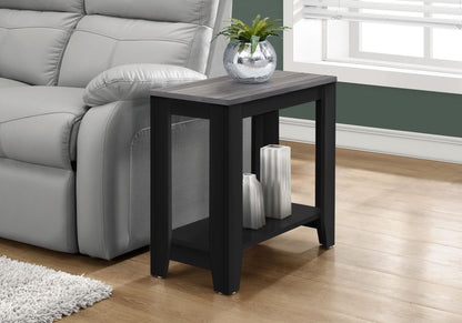 An Accent Table, Side Transitional with elegant design in black and gray stands by a gray sofa. It displays a round glass vase with a green plant on top and two white decorative vases on its lower shelf.