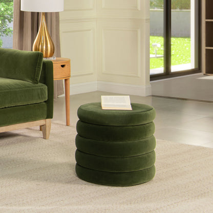 The Fuji Oversized Round Storage Ottoman, upholstered in green velvet with a stacked ring design, sits on a beige rug beside a matching sofa. An open book rests on top as sunlight pours through large glass doors in the modern living room.