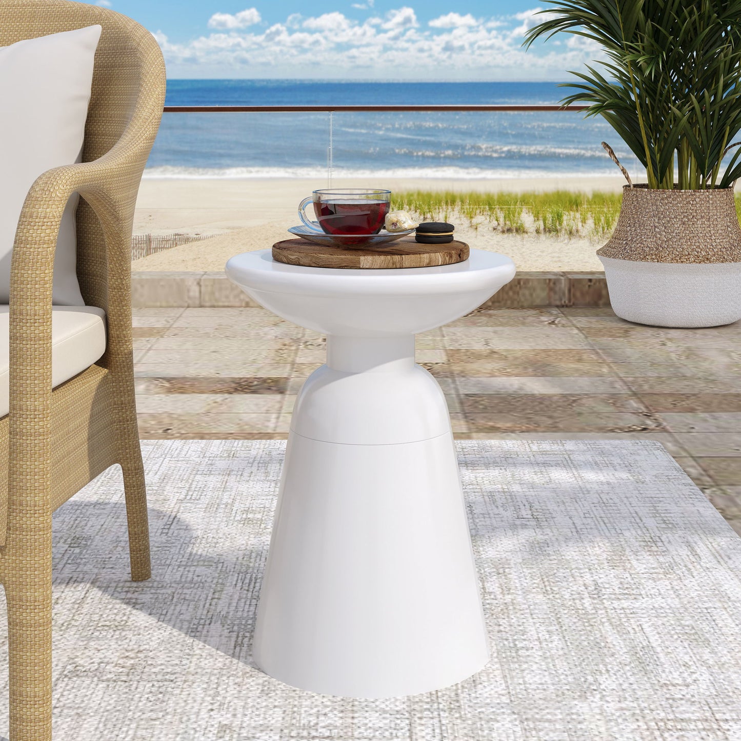 The Outdoor Modern Metal End Large Table, topped with a glass teacup and saucer on a wooden tray, stands near a wicker chair on the patio with a scenic beach view of sand, sea, and blue sky.