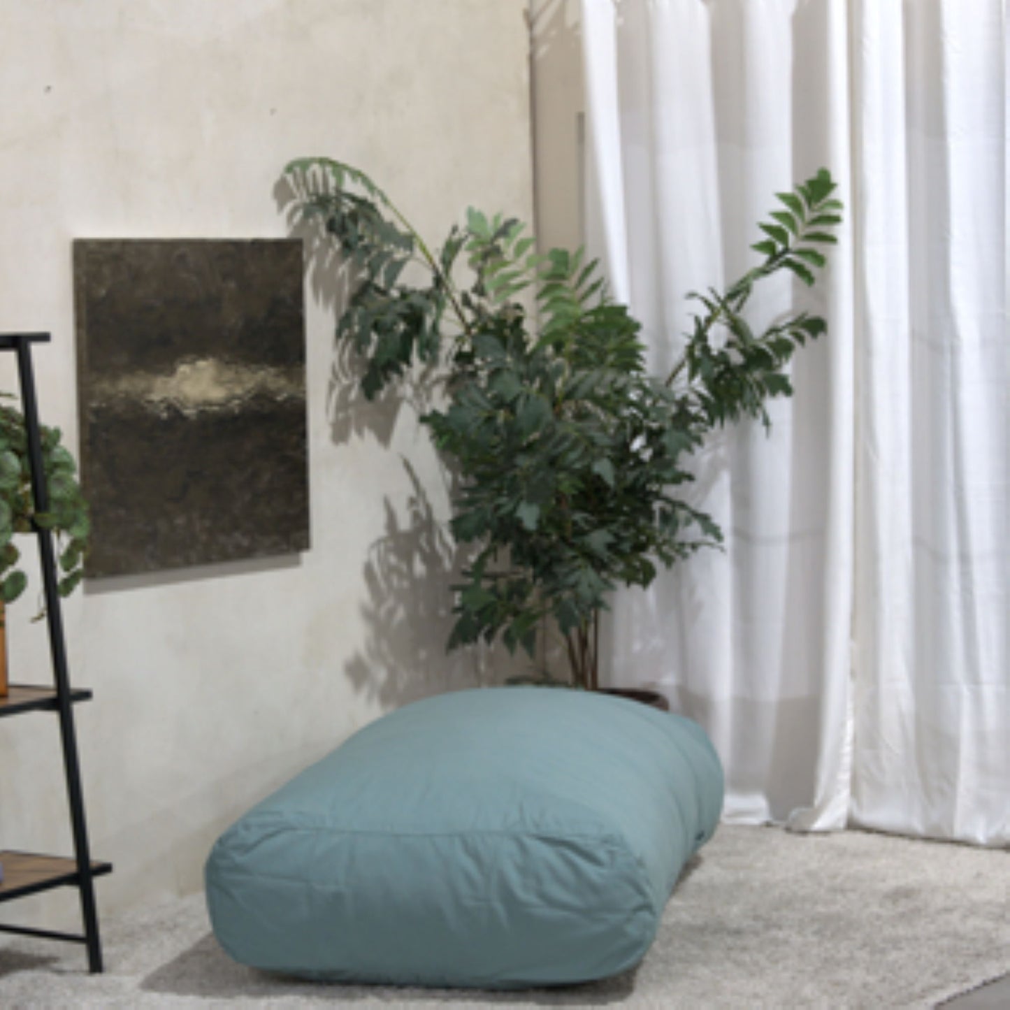 A cozy corner featuring the Kasper Water Resistant Lounger Bean Bag in blue, a leafy green plant, a small painting, white curtains, and part of a black metal shelf with wooden shelves.