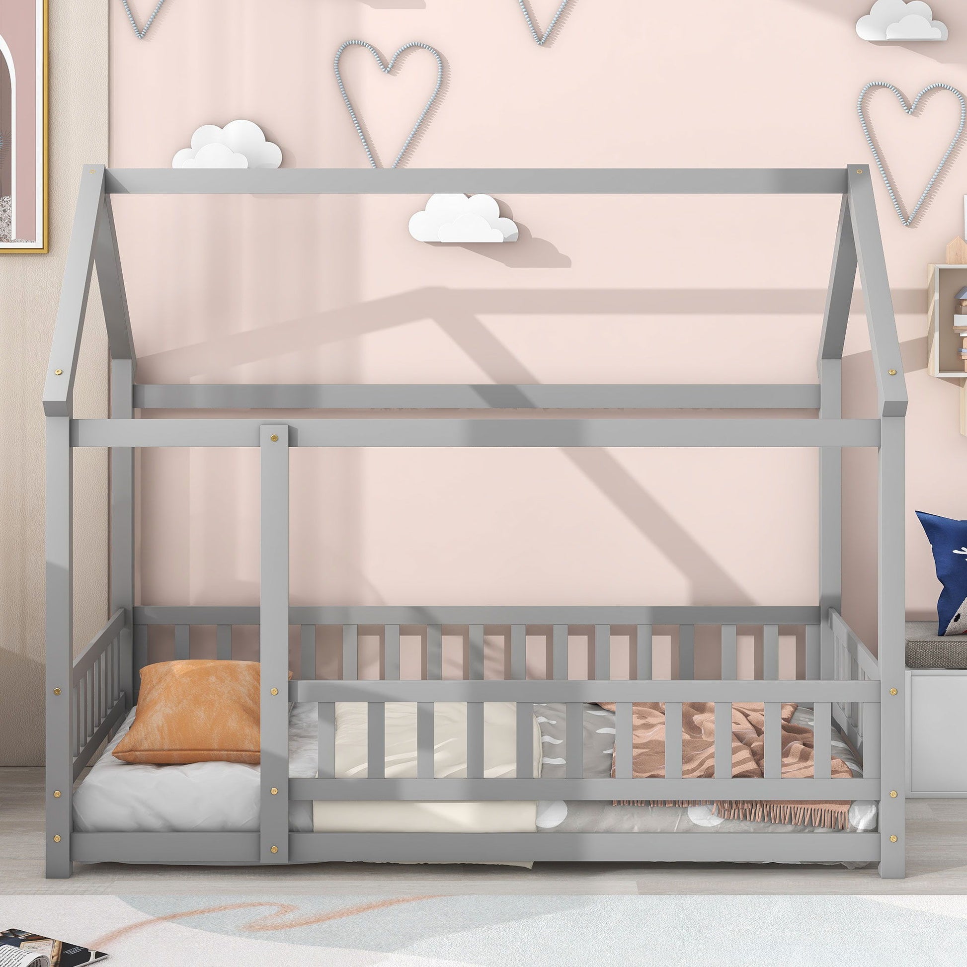 The Floor Wooden Bed With House Roof Frame and fence guardrails is shown in gray, styled with pillows and blankets. The backdrop has a pink wall decorated with heart and cloud-shaped wall art.