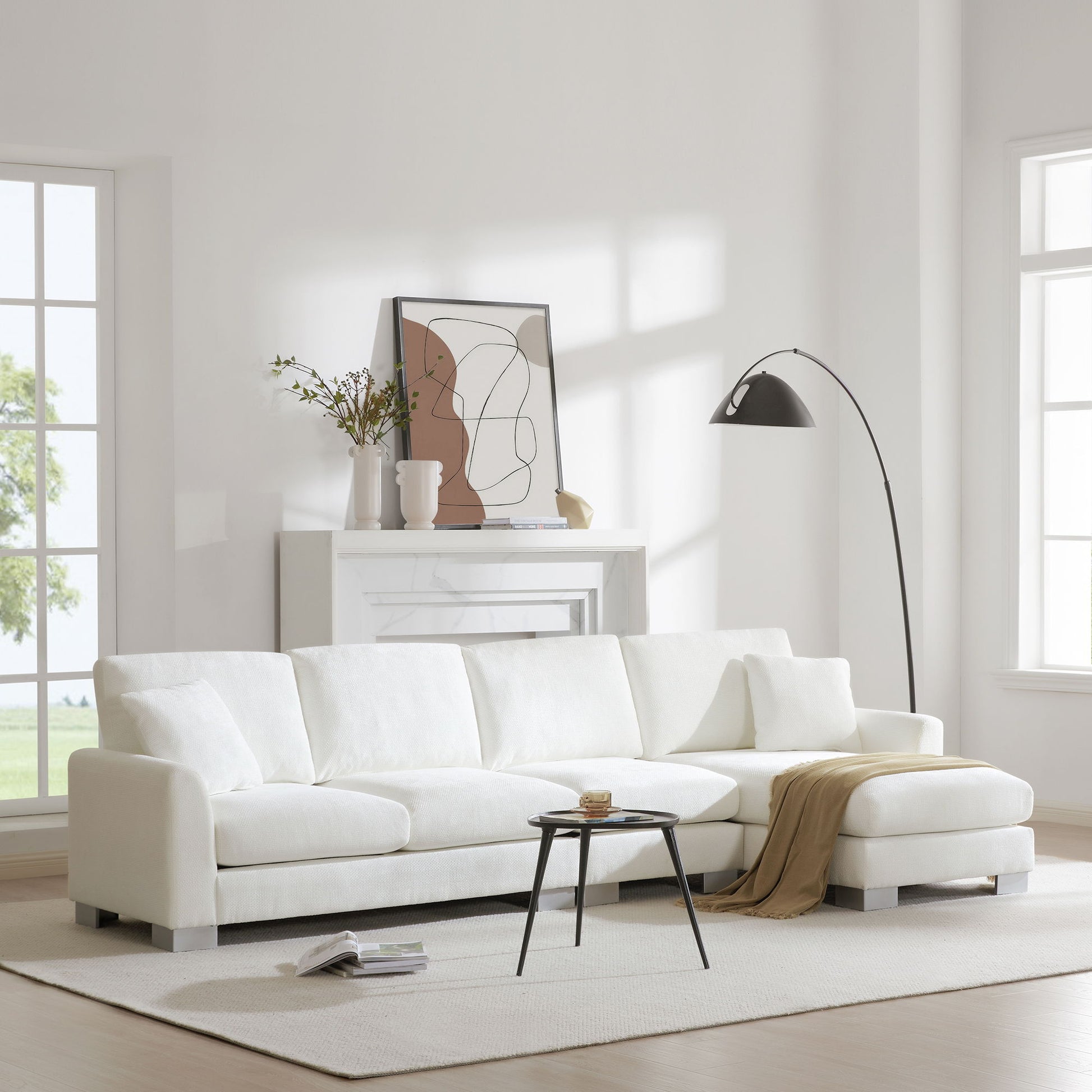A bright, modern living room featuring a Modern Oversized Sectional Sofa, L-shaped with chaise and two pillows, paired with a round black coffee table, beige throw, abstract mantel art, vases, a black floor lamp, and large windows filling the space with light.