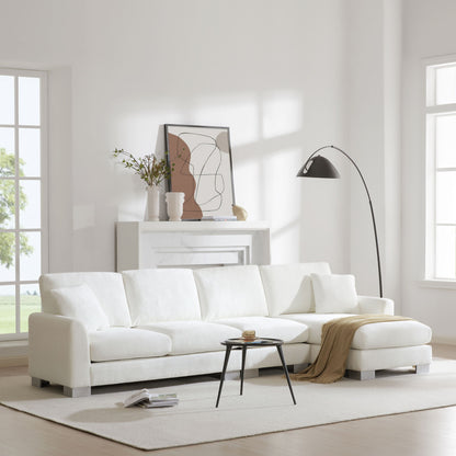 A bright, modern living room featuring a Modern Oversized Sectional Sofa, L-shaped with chaise and two pillows, paired with a round black coffee table, beige throw, abstract mantel art, vases, a black floor lamp, and large windows filling the space with light.