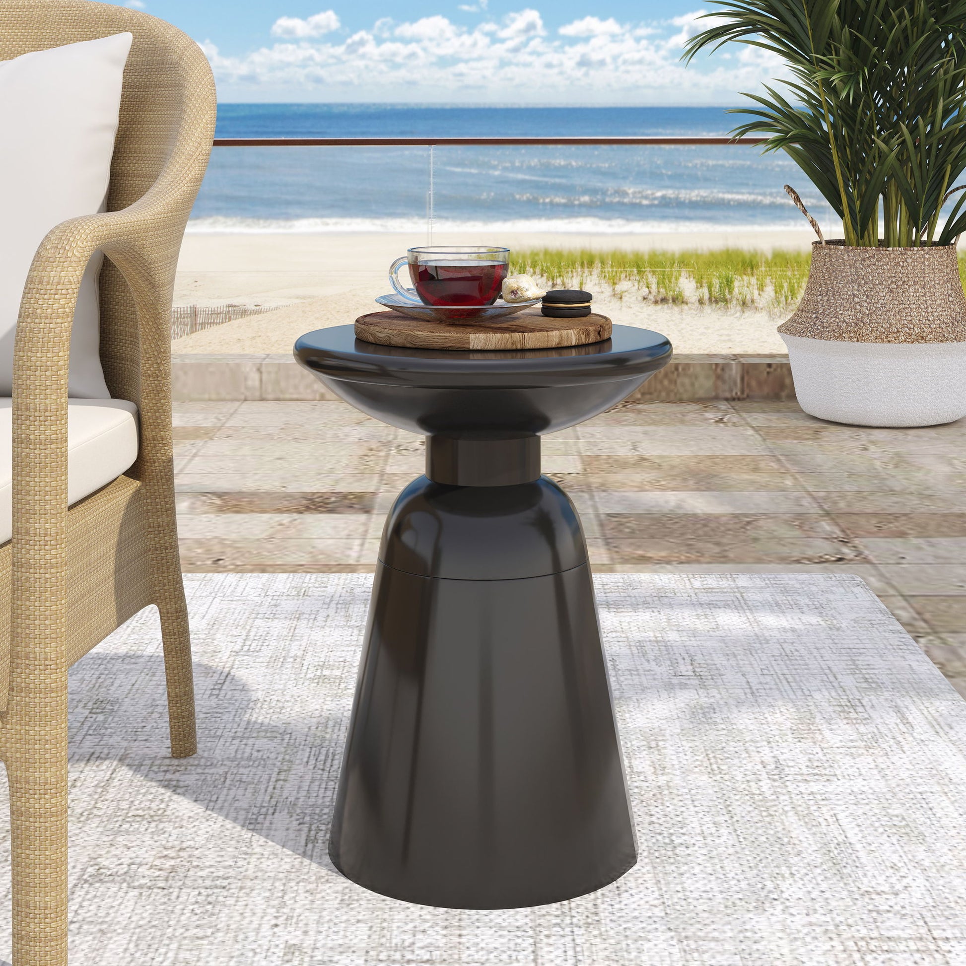 The Outdoor Modern Metal End Large Table, in sleek black, sits on the patio with a cup of tea and wooden tray. A wicker chair, lush plant, and a sandy beach with blue ocean and sky are seen in the background.