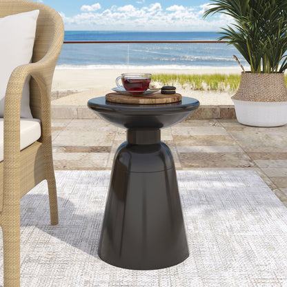 The Outdoor Modern Metal End Large Table, in sleek black, sits on the patio with a cup of tea and wooden tray. A wicker chair, lush plant, and a sandy beach with blue ocean and sky are seen in the background.